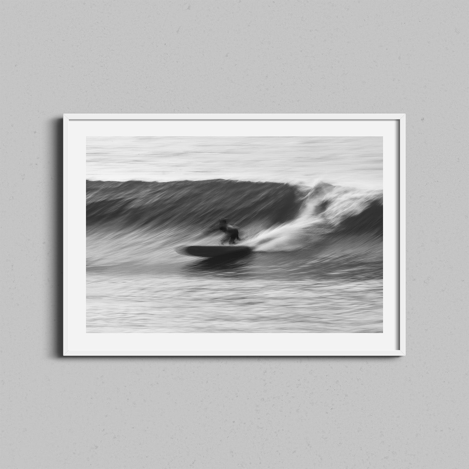 A framed black-and-white photograph of a surfer surfing a wave. The wall art is framed in a a white wooden frame.