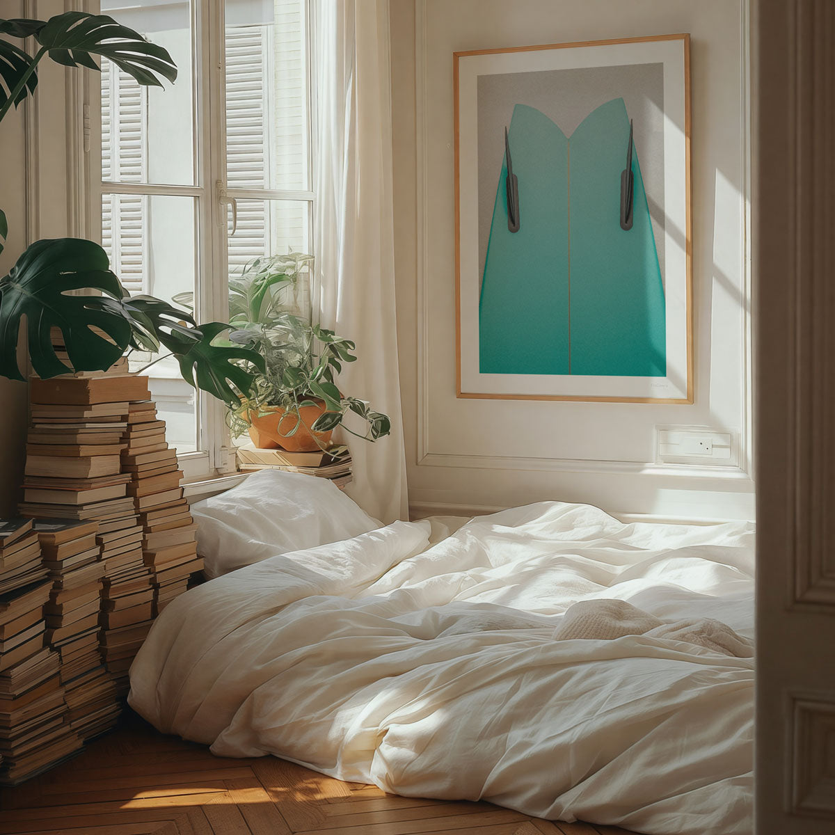 A framed coastal photograph featuring a green surfboard in a natural wooden frame. The art hangs on a wall in a boho-chic bedroom with many books stacked against a wall.