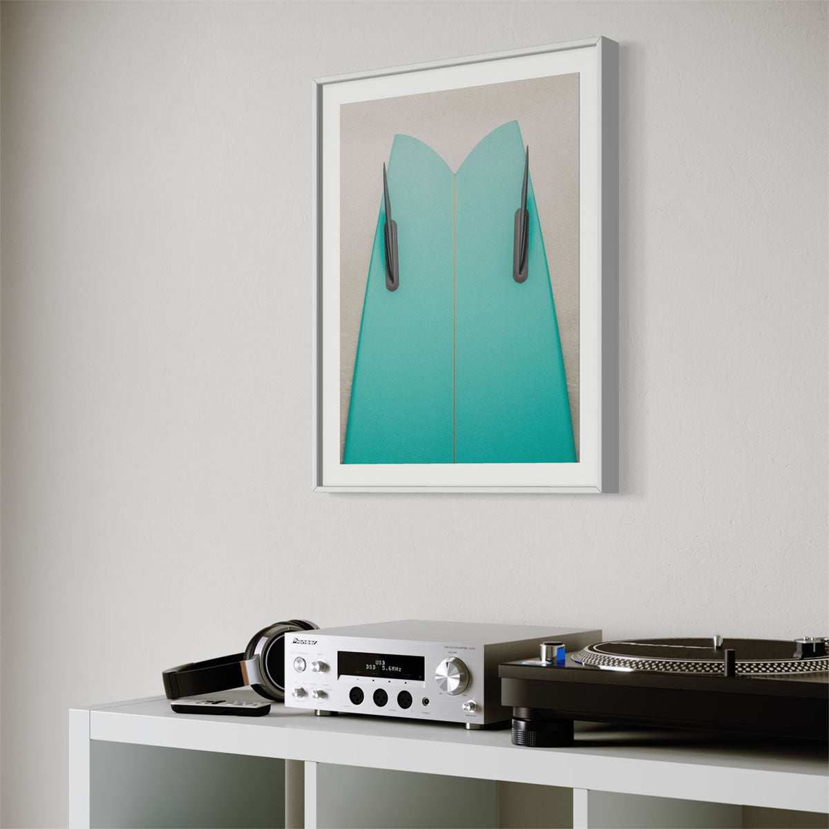 A framed coastal photograph featuring a green surfboard. The art hangs on a wall in a modern living room with a record player on a shelf.