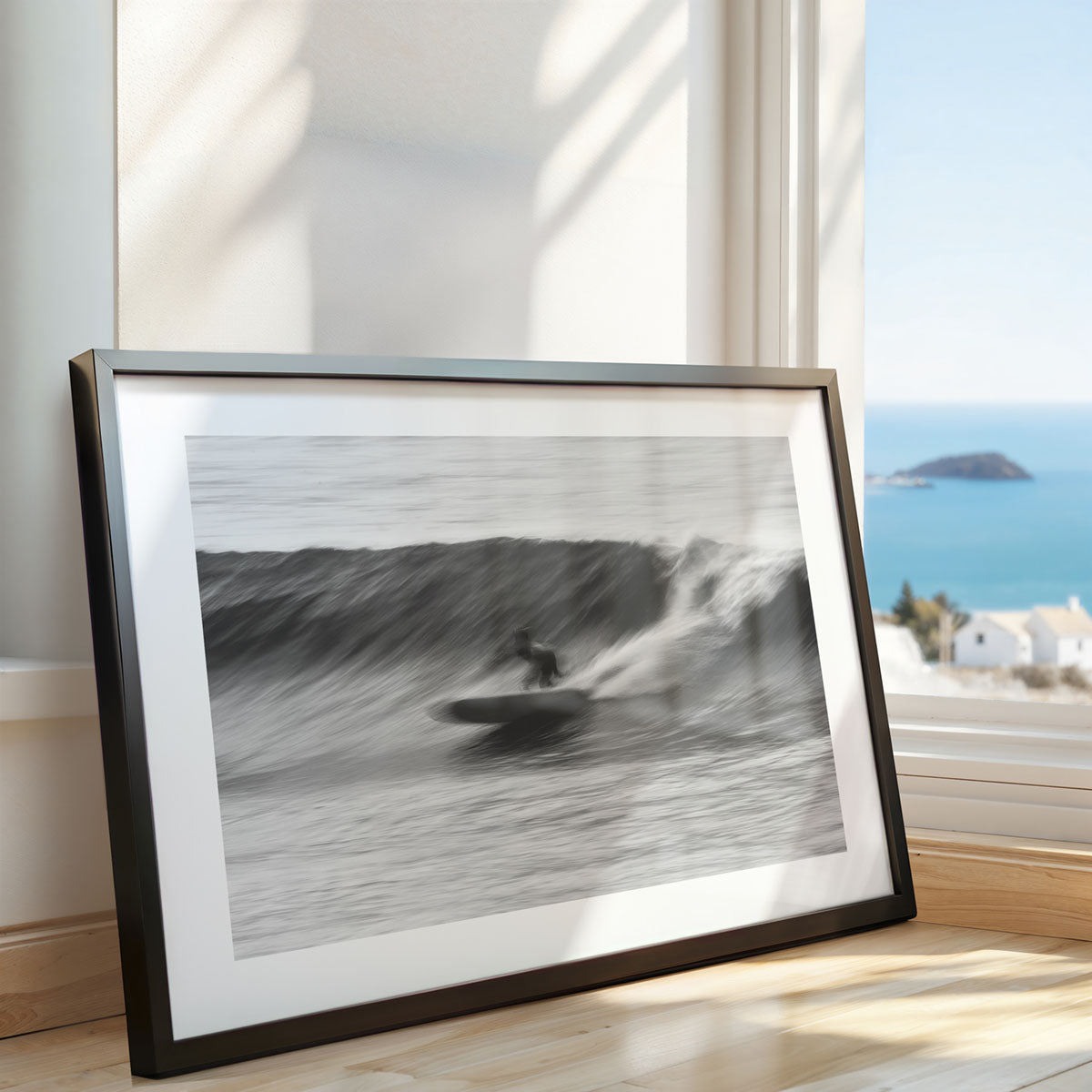 A framed black-and-white photograph of a  surfer captured surfing a wave. The wall art sits next to a window with a coastal view. 