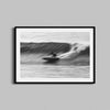 A framed black-and-white photograph of a  surfer surfing a wave. The wall art is framed in a a black wooden frame.