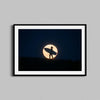 A surfer holds a surfboard against the backdrop of a full moon in this black- framed photography print.