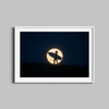 A surfer holds a surfboard against the backdrop of a full moon in this framed photography print.